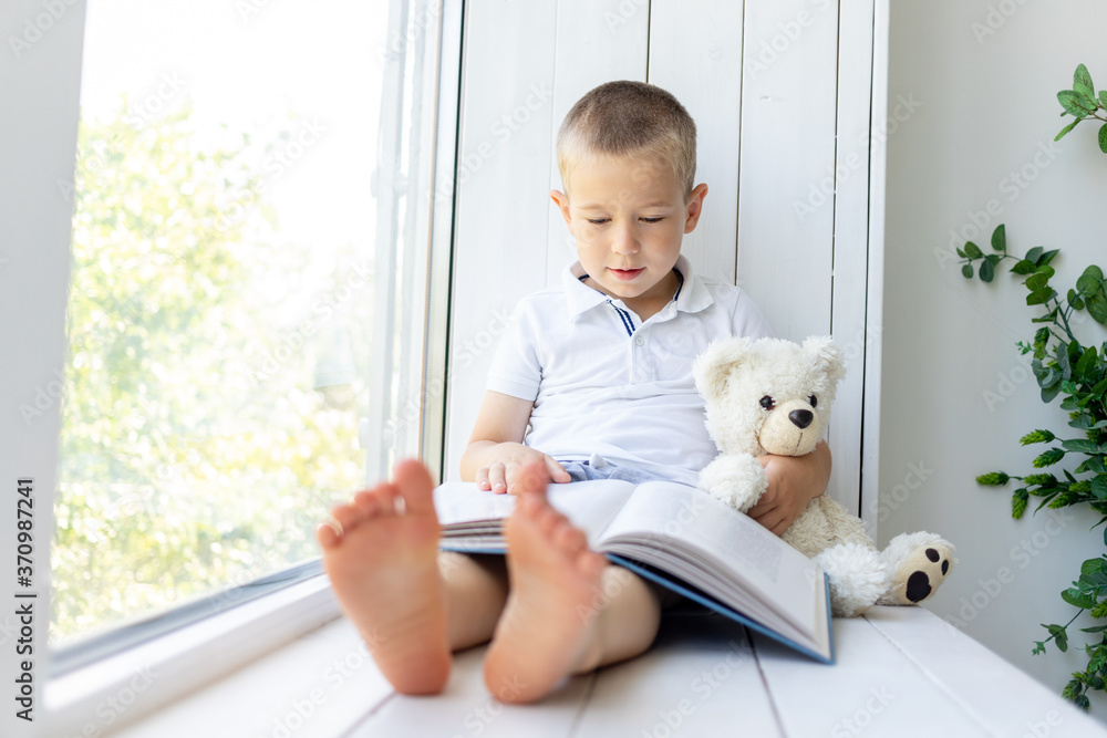 a small boy sits on the window with a book and a soft bear a small boy sits on the window with a book and a soft bear