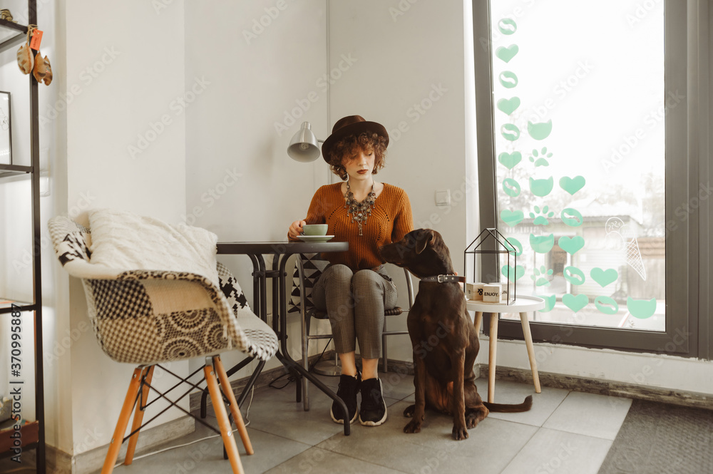 Beautiful girl in vintage clothes spends time with a dog in a cozy cafe, holds a cup of coffee in her hand and looks at a pet. Stylish lady is resting in a pet friendly cafe with a pet.
