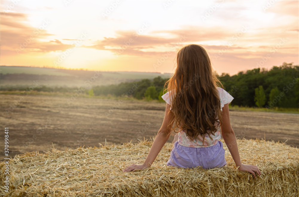 Adorable little girl child sitting on a hay rolls in a wheat field at sunset Adorable little girl child sitting on a hay rolls in a wheat field at sunset