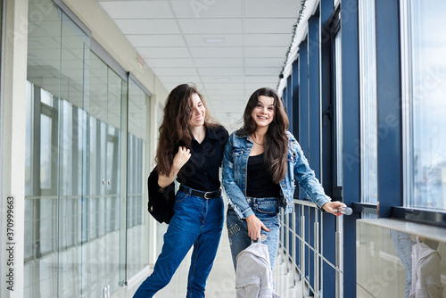Two young brunette girls  standing in light airport hallway with huge windows wearing casual jeans clothes  holding white and black backpacks.Girlfriends  waiting for flight  smiling. Safe air travel.
