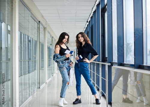 Two young brunette girls  standing in light airport hallway with huge windows  wearing casual jeans clothes  holding international passports and boarding passes tickets. Girlfriends traveling by air.