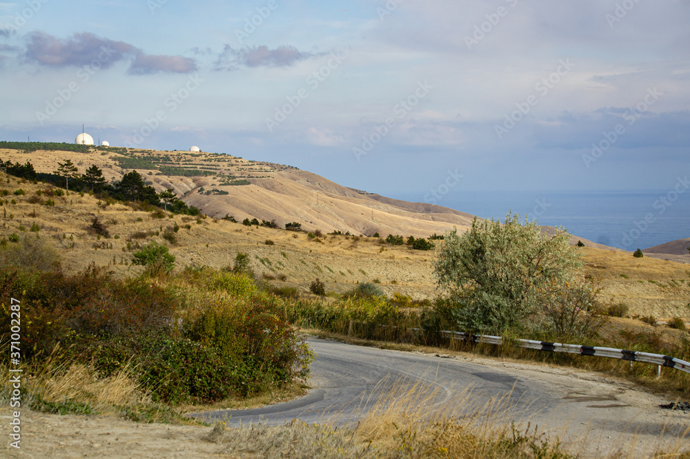 Winding road in mountains. Panoramic view of mountains of Black Sea coast of Crimea from height of mountain range between Feodosia and Koktebel. In background are white balls of Crimean observatory. Winding road in mountains. Panoramic view of mountains of Black Sea coast of Crimea from height of mountain range between Feodosia and Koktebel. In background are white balls of Crimean observatory.