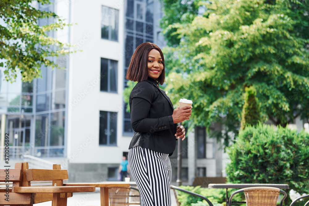 Coffee break. Young afro american woman in fashionable clothes outdoors in the city near green trees and against business building