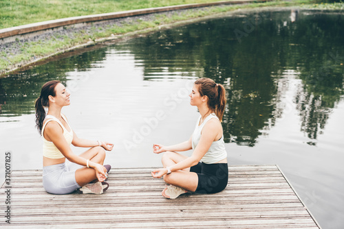 Attractive tranquil young women in sportswear do yoga exercises sitting in lotus poses on wooden pier near calm pound on nice summer day. © Mariia Korneeva