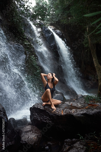 beautiful brunette sits against the background of a waterfall and washes under it.