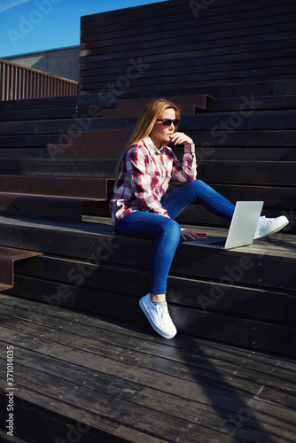 Shot of caucasian college student studying with laptop computer at campus, attractive young woman using laptop sitting on wooden staircase enjoying sunny day outdoors, charming girl