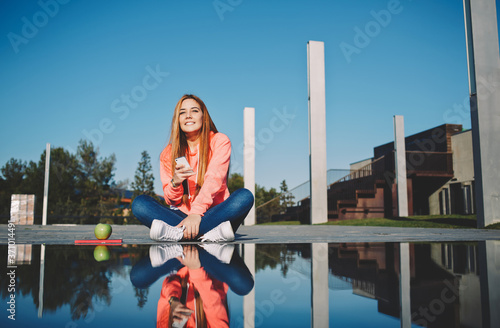 Gorgeous young woman holding her smart phone sitting at beautiful park on sunny day, attractive girl sitting next to the water with amazing reflection of her self, female student using cell phone