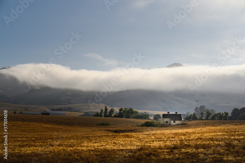 Farmhouse in early morning in golden light with golden field and low hanging clouds with mountains in the background