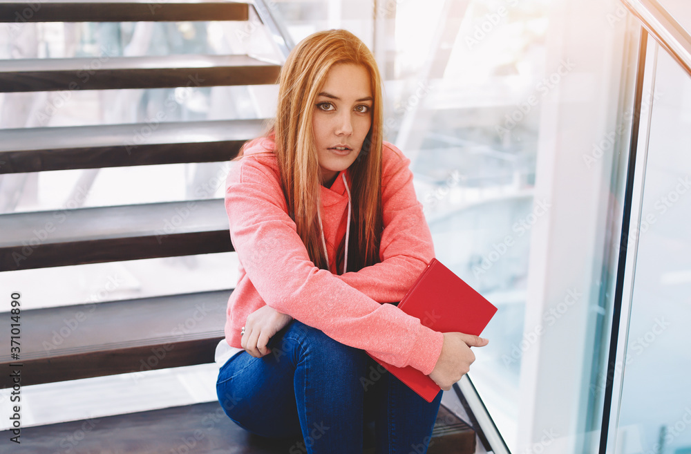 Portrait of cute young college student holding exercise book while sitting on the stairs of college hallway, female hipster in trendy clothes girl resting after preparation for lessons at school Portrait of cute young college student holding exercise book while sitting on the stairs of college hallway, female hipster in trendy clothes girl resting after preparation for lessons at school
