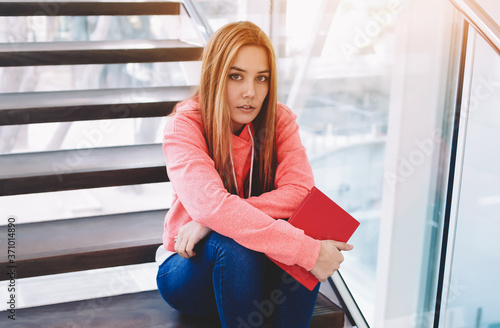 Portrait of cute young college student holding exercise book while sitting on the stairs of college hallway, female hipster in trendy clothes girl resting after preparation for lessons at school