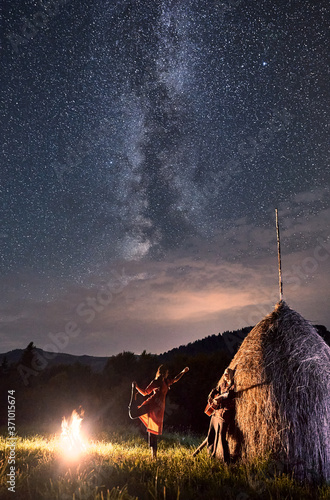 Romantic couple having a rest in the mountains, a girl dancing next to the fire and a guy playing the guitar leaned on rick of dry hay under incredible beautiful night sky full of stars and Milky way