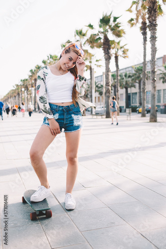 Full length portrait of young happy glamorous woman posing with her longboard while standing on the street in sunny day, smiling charming female skateboarder enjoy rest after riding on penny board