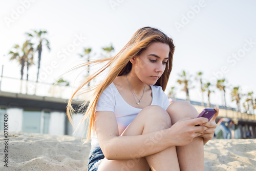 Gorgeous female enjoying recreation time while focused chatting on her mobile phone while sitting on the beach in summer day, pretty young woman holding smart phone in the hand while resting outdoors