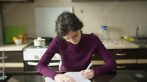Cute student girl makes notes in a notebook, sitting at the kitchen table