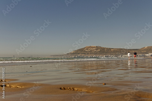 Agadir, Morocco.Feb.9,2019:Beach with tourist walking and Moroccan motto on the mountain. Writing on the hillside meaning, God, Country, King. People walking on the beach footsteps in the sand. © Erik