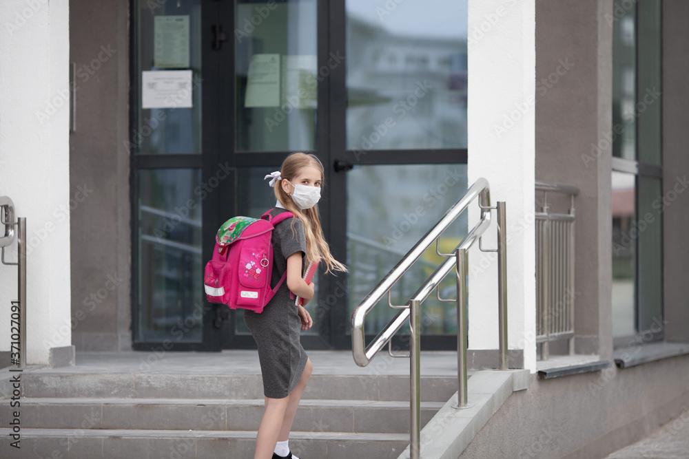 A schoolgirl wearing medical face mask and carrying a pink backpack goes up the stairs of her school, looking back to her parents. Back to school. Protection from virus for school.