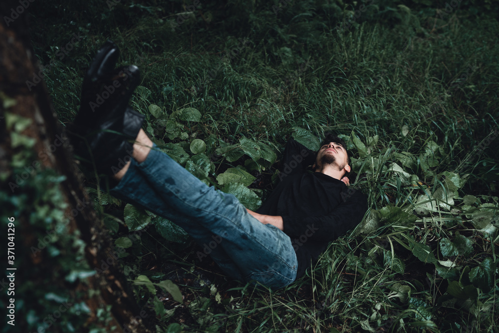 Handsome man in the forest stretches his feet to a tree and lies on the wet grass