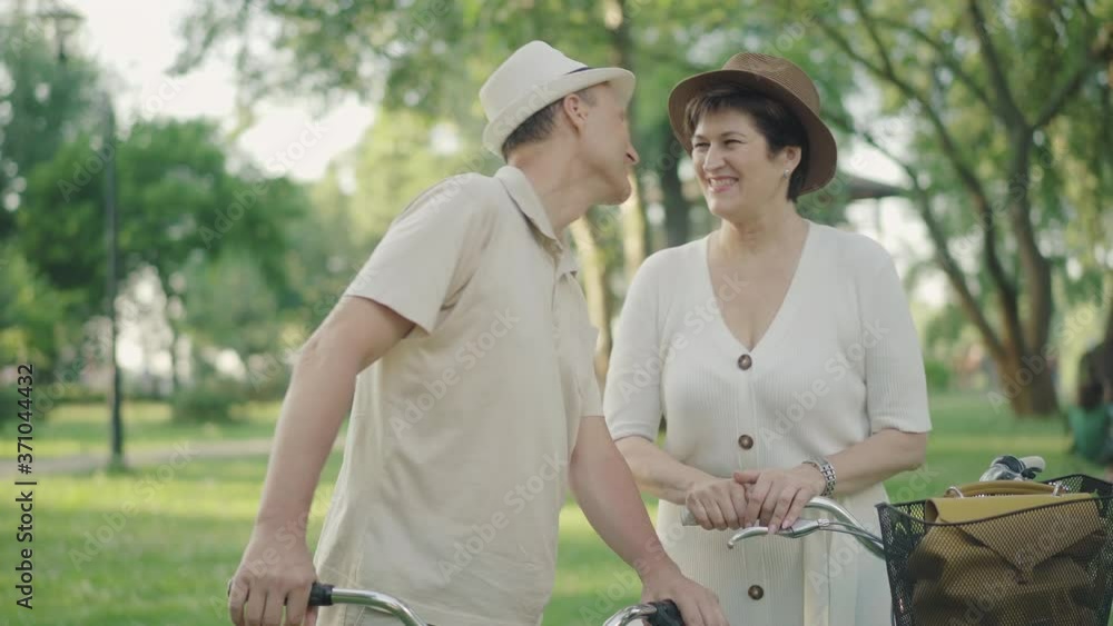 Handsome mid-adult man kissing wife on cheek and talking with loving spouse. Portrait of happy mid-adult Caucasian couple enjoying sunny summer day in park. Positive family dating outdoors. Handsome mid-adult man kissing wife on cheek and talking with loving spouse. Portrait of happy mid-adult Caucasian couple enjoying sunny summer day in park. Positive family dating outdoors.