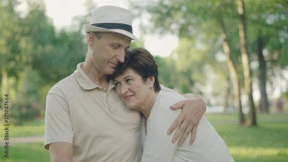 Elegant mid-adult husband hugging beautiful wife and kissing her head. Portrait of happy loving Caucasian family resting in sunny park on weekends. Cheerful couple enjoying summer day outdoors. Elegant mid-adult husband hugging beautiful wife and kissing her head. Portrait of happy loving Caucasian family resting in sunny park on weekends. Cheerful couple enjoying summer day outdoors.