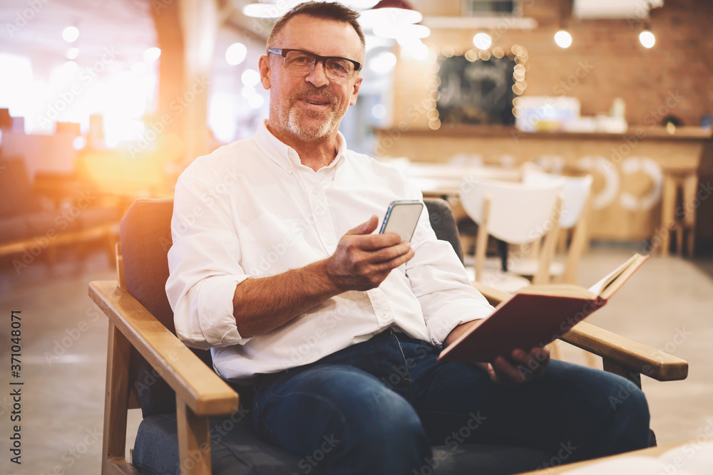 Portrait of cheerful male executive holding gadget while chatting with assistant while resting with favorite book after hard working day sitting in cozy interior cafe using free wireless connection