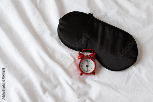 Silk black sleep mask, red clock, showing 6 o'clock on white rumpled sheets. Top view, flat lay. Horizontal. Concept of rest, awakening, sleep, good morning. For social media, blog. Minimal style