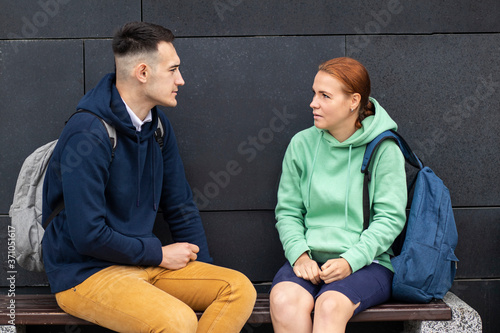 Couple of young university or college students with backpacks, boy chatting with a girl, having a conversation. Man and woman sitting on a bench outdoors in campus, talking, looking to each other. photo