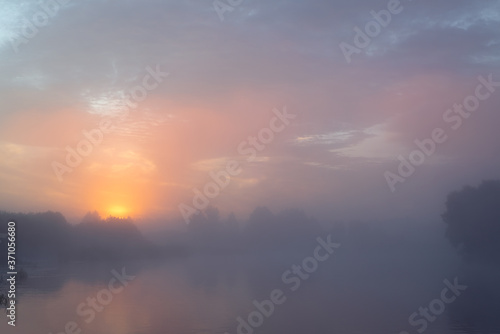 Tranquil morning landscape at sunrise. Heavy fog over the river. Dawn illuminates and makes the clouds colorful.
