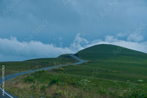 the roadbed is uneven, winding, passes through mountains and cliffs. Clouds are dark, low, with rain. A thick white fog is spreading along the road.