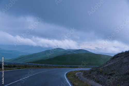 the roadbed is uneven, winding, passes through mountains and cliffs. Clouds are dark, low, with rain. A thick white fog is spreading along the road.