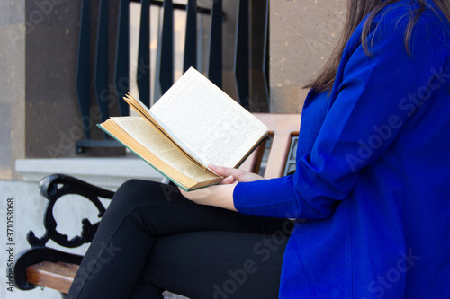 A woman in a blue jacket is reading a book sitting on a bench photo