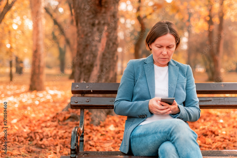 Woman typing text on mobile phone on autumn park bench Woman typing text on mobile phone on autumn park bench