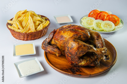 Delicious whole grilled chicken with french fries, salad and creams on wooden plate. Peruvian food "pollo a la brasa con papas fritas". White background.