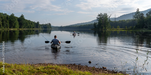 A young man and woman with backs facing the camera on kayaks on mountain lake early in the morning.   Oars are in motion with ripples on the water.    A back drop of mountains and blue sky with clouds
