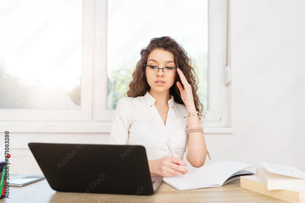 Attractive bussineswomen with curly hair wearing glasses, looking at laptop and writing in the book at workplace. Attractive bussineswomen with curly hair wearing glasses, looking at laptop and writing in the book at workplace.