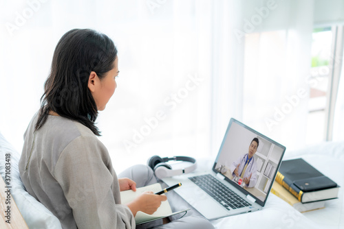 Back view of Asian woman making video call with her doctor with her feeling sick on laptop in bedroom for online healthcare digital technology service consultation while staying at home.