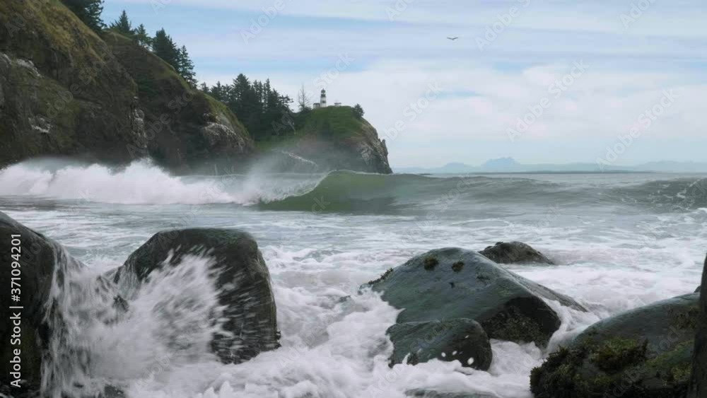 Rugged coast ocean shore multiple big curling waves crashing hard on rocks with white lighthouse on top cliff in background at Cape Disappointment State Park pacific northwest Washington shot Rugged coast ocean shore multiple big curling waves crashing hard on rocks with white lighthouse on top cliff in background at Cape Disappointment State Park pacific northwest Washington shot