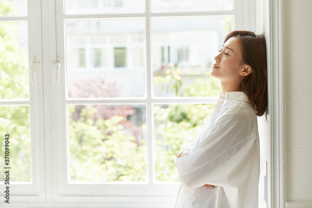 young asian woman staying at home standing by the window arms crossed eyes closed