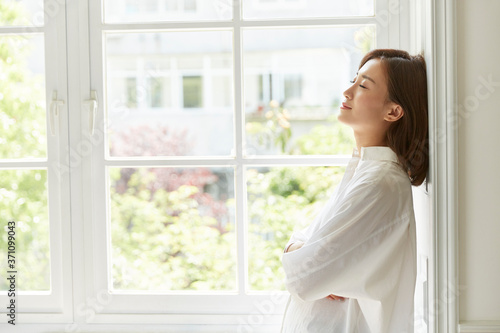 young asian woman staying at home standing by the window arms crossed eyes closed