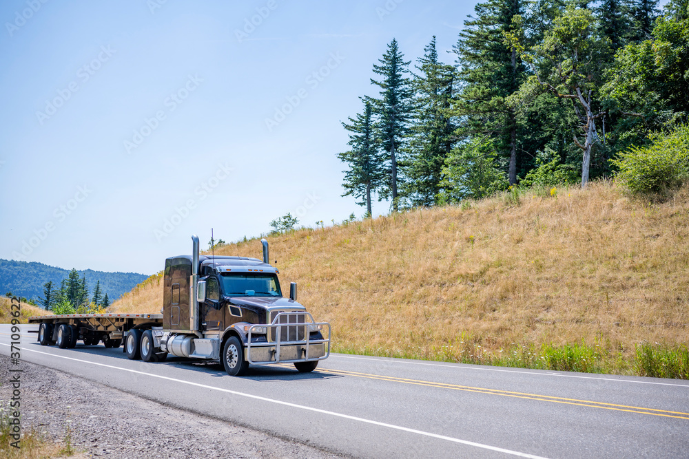 Powerful big rig black semi truck with grille guard going up hill with empty flat bed semi trailer in gorgeous Columbia Gorge area