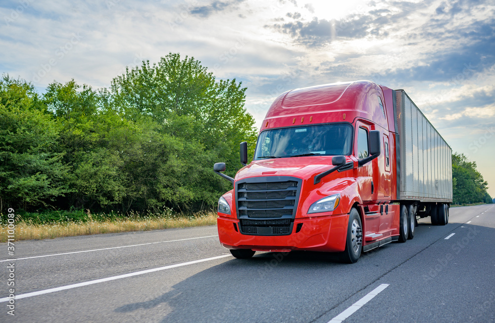 Red big rig long haul semi truck with black grille transporting cargo in dry van semi trailer running on the wide highway road Red big rig long haul semi truck with black grille transporting cargo in dry van semi trailer running on the wide highway road