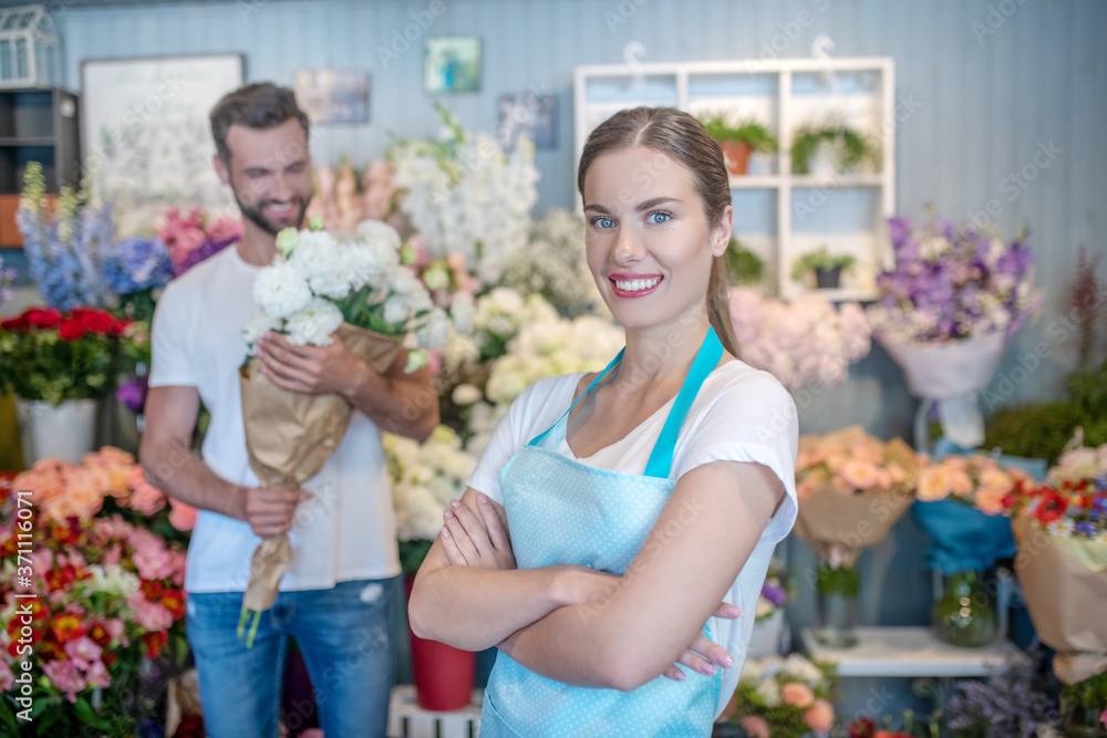 Charming female florist crossing her arms, male standing with bouquet of white flowers behind her