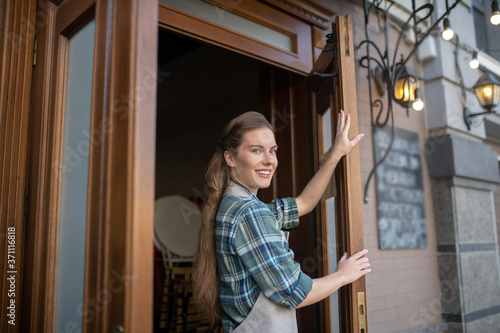 Smiling young waitress standing at the entrance, holding doors