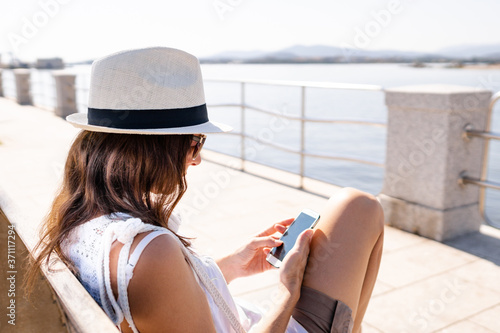 Caucasian young woman with white hat sitting on a bench of a sidewalk by the sea using her smartphone - Single woman outdoor connected with new social technology © Robby Fontanesi