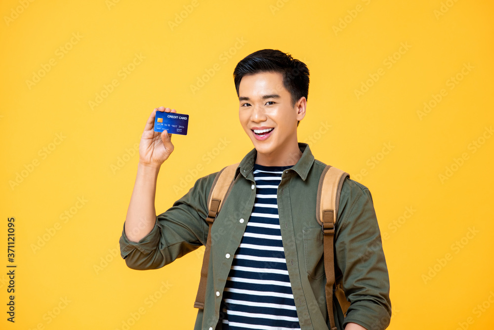 Portrait of a smiling young Asian backpacker tourist man showing credit card in isolated studio yellow background Portrait of a smiling young Asian backpacker tourist man showing credit card in isolated studio yellow background