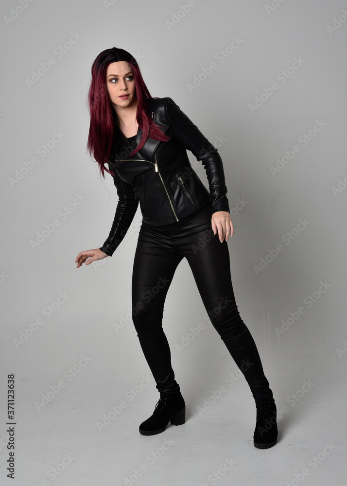 Full length portrait of girl with red hair wearing black leather jacket, pants and boots. Standing pose, isolated against a grey studio background. Full length portrait of girl with red hair wearing black leather jacket, pants and boots. Standing pose, isolated against a grey studio background.