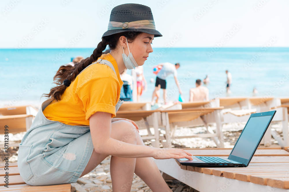 Freelance. Portrait of woman wearing medical mask on her face, sitting on a chaise longue and typing on a laptop. In the background, the beach and the sea. The concept of remote work during a pandemic