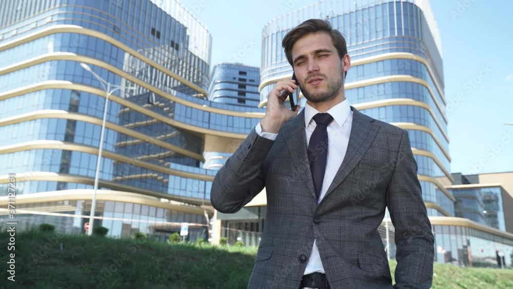 Businessman, top manager in a suit stands near a glass skyscraper and talking on a mobile phone, young businessman manager near the modern office. Businessman, top manager in a suit stands near a glass skyscraper and talking on a mobile phone, young businessman manager near the modern office.