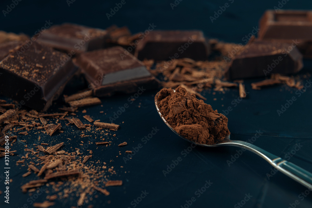 Pieces of chocolate, chocolate shavings and spoon with cocoa powder on dark blue textured background.