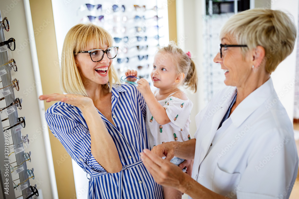 Happy mother with her cute daughter choosing glasses in optics store