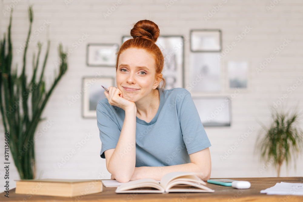 Tired bored redhead young woman student sitting at a table with textbooks and a workbook, looking at camera. Girl student preparing for college exams. Stressed female during an exams on the classroom Tired bored redhead young woman student sitting at a table with textbooks and a workbook, looking at camera. Girl student preparing for college exams. Stressed female during an exams on the classroom
